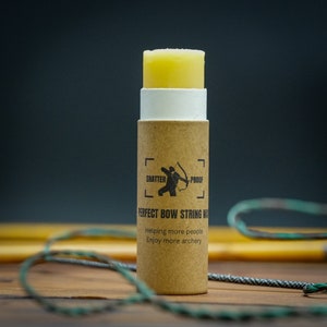 May include: A cylindrical container of bow string wax. The wax is yellow and sits atop a white and brown cardboard tube. The tube has the words "SHATTER PROOF" and "PERFECT BOW STRING WAX" printed on it, along with a graphic of an archer.
