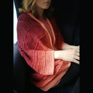 May include: A crocheted cardigan with a gradient of red, orange and peach colours. The cardigan has a loose fit and long sleeves.