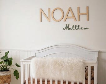 May include: A white wooden crib with a fluffy cream-colored blanket and a rust-colored patterned sheet. Above the crib, the name NOAH is displayed in large wooden letters, with the name Matthew in a smaller, dark green cursive script.