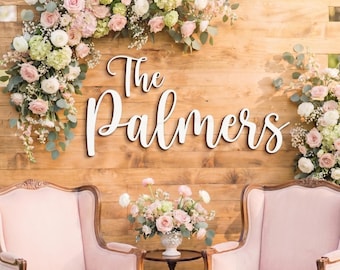 May include: A wooden backdrop featuring the white script text The Palmers, adorned with a large floral arrangement of pink roses, white ranunculus, and green hydrangeas. Two light pink upholstered armchairs flank a small table with a floral centerpiece.