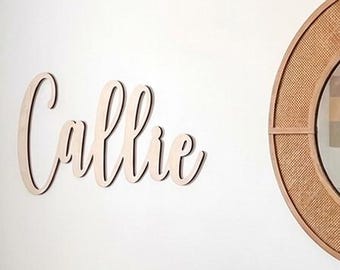 May include: A round mirror with a light brown wooden frame and woven cane detailing, featuring a reflection of a pastel rainbow. To the left, a wooden script sign reads Callie. Below, a black changing pad and a pink ribbed storage box.