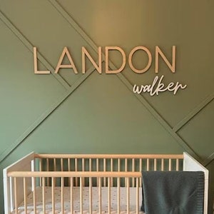 Peut inclure: Une pancarte en bois avec le nom "LANDON walker" en bois clair, montée sur un mur vert avec un lit bébé blanc et un jouet en peluche en forme de girafe au premier plan.