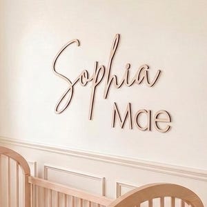May include: A light-colored wooden crib with arched ends and a pink sheet, featuring a patterned blanket draped over the side. Mounted on the wall above the crib is a wooden sign with the name Sophia Mae written in a cursive and print font.