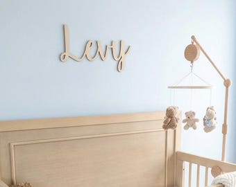 May include: A light wood crib with a white mattress featuring a brown teddy bear print. A wooden mobile with three hanging teddy bears stands beside the crib. A cream-colored chevron knit blanket is draped over the side. The name Levy is mounted on the wall in wooden script lettering.