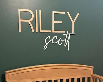 May include: A wooden crib with a light brown finish containing a crocheted blanket featuring a granny square pattern in dark green, brown, and cream. Above the crib, the name RILEY in block letters and scott in script are mounted on the wall.