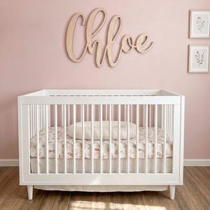 K&ouml;nnte beinhalten: Ein wei&szlig;es Babybett mit einem floral gemusterten Laken und einem Kissen. &Uuml;ber dem Bett steht in Schreibschrift der Name "Chloe" auf einem Holzschild. Die Wand ist zartrosa gestrichen, und es gibt zwei gerahmte botanische Drucke.