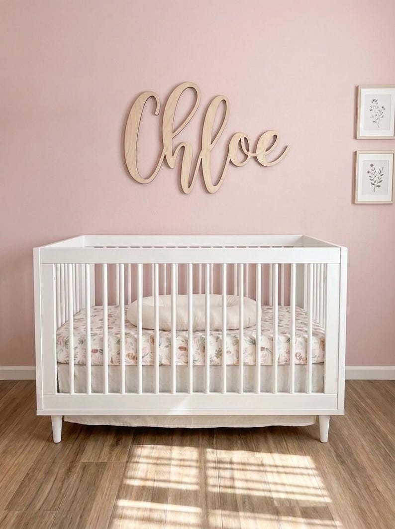 K&ouml;nnte beinhalten: Ein wei&szlig;es Babybett mit einem floral gemusterten Laken und einem Kissen. &Uuml;ber dem Bett steht in Schreibschrift der Name "Chloe" auf einem Holzschild. Die Wand ist zartrosa gestrichen, und es gibt zwei gerahmte botanische Drucke.