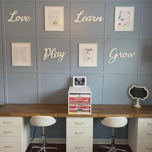 K&ouml;nnte beinhalten: Ein Homeoffice mit hellblauer Wand, gerahmten Kunstwerken und wei&szlig;en Holzbuchstaben, die "Love", "Learn", "Play" und "Grow" buchstabieren. Ein Schreibtisch mit wei&szlig;en Schubladen und einer Holzplatte hat zwei wei&szlig;e St&uuml;hle. Eine kleine Tafel und eine Digitaluhr sind ebenfalls sichtbar.