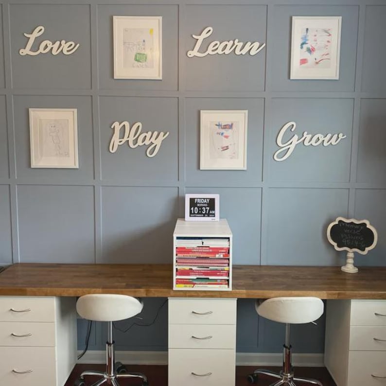 K&ouml;nnte beinhalten: Ein Homeoffice mit hellblauer Wand, gerahmten Kunstwerken und wei&szlig;en Holzbuchstaben, die "Love", "Learn", "Play" und "Grow" buchstabieren. Ein Schreibtisch mit wei&szlig;en Schubladen und einer Holzplatte hat zwei wei&szlig;e St&uuml;hle. Eine kleine Tafel und eine Digitaluhr sind ebenfalls sichtbar.