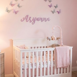 K&ouml;nnte beinhalten: Ein wei&szlig;es Babybett mit einem rosa gebl&uuml;mten Laken, einer rosa Decke und einem Mobile mit Stofftieren. Die rosa Wand ist mit Schmetterlingsdekorationen und dem Namen "Aryanna" in Schreibschrift versehen.