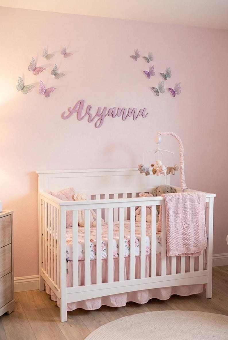 K&ouml;nnte beinhalten: Ein wei&szlig;es Babybett mit einem rosa gebl&uuml;mten Laken, einer rosa Decke und einem Mobile mit Stofftieren. Die rosa Wand ist mit Schmetterlingsdekorationen und dem Namen "Aryanna" in Schreibschrift versehen.