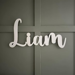 K&ouml;nnte beinhalten: Wei&szlig;es Holzschild mit dem Namen "Liam" in Schreibschrift. Das Schild ist an einer grauen Wand mit einem Rastermuster befestigt. Die Buchstaben sind sauber und modern und schaffen ein schlichtes, aber elegantes Design.