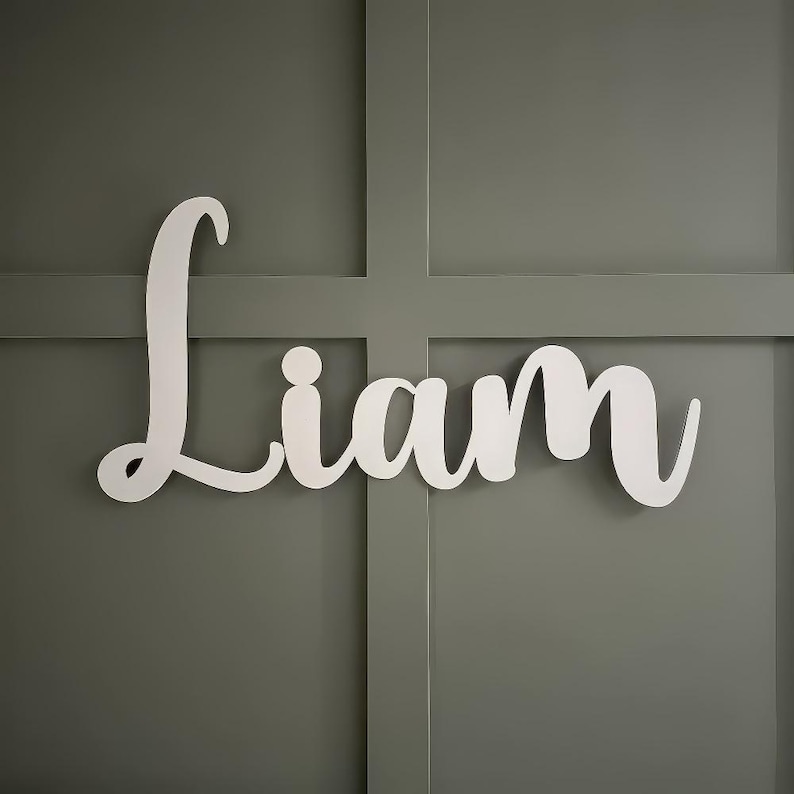 K&ouml;nnte beinhalten: Wei&szlig;es Holzschild mit dem Namen "Liam" in Schreibschrift. Das Schild ist an einer grauen Wand mit einem Rastermuster befestigt. Die Buchstaben sind sauber und modern und schaffen ein schlichtes, aber elegantes Design.