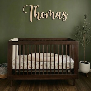 K&ouml;nnte beinhalten: Ein dunkelbraunes Holzbettchen mit einer gemusterten Matratze und einem beigen Kissen. &Uuml;ber dem Bettchen ist der Name "Thomas" in h&ouml;lzernen Schreibschriftbuchstaben dargestellt. Der Hintergrund ist eine dunkelgr&uuml;ne Wand, mit einer kleinen Pflanze in einem wei&szlig;en Topf rechts.