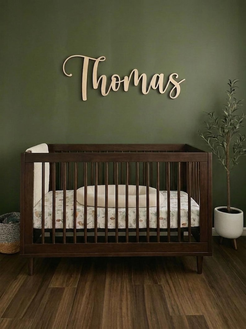 K&ouml;nnte beinhalten: Ein dunkelbraunes Holzbettchen mit einer gemusterten Matratze und einem beigen Kissen. &Uuml;ber dem Bettchen ist der Name "Thomas" in h&ouml;lzernen Schreibschriftbuchstaben dargestellt. Der Hintergrund ist eine dunkelgr&uuml;ne Wand, mit einer kleinen Pflanze in einem wei&szlig;en Topf rechts.