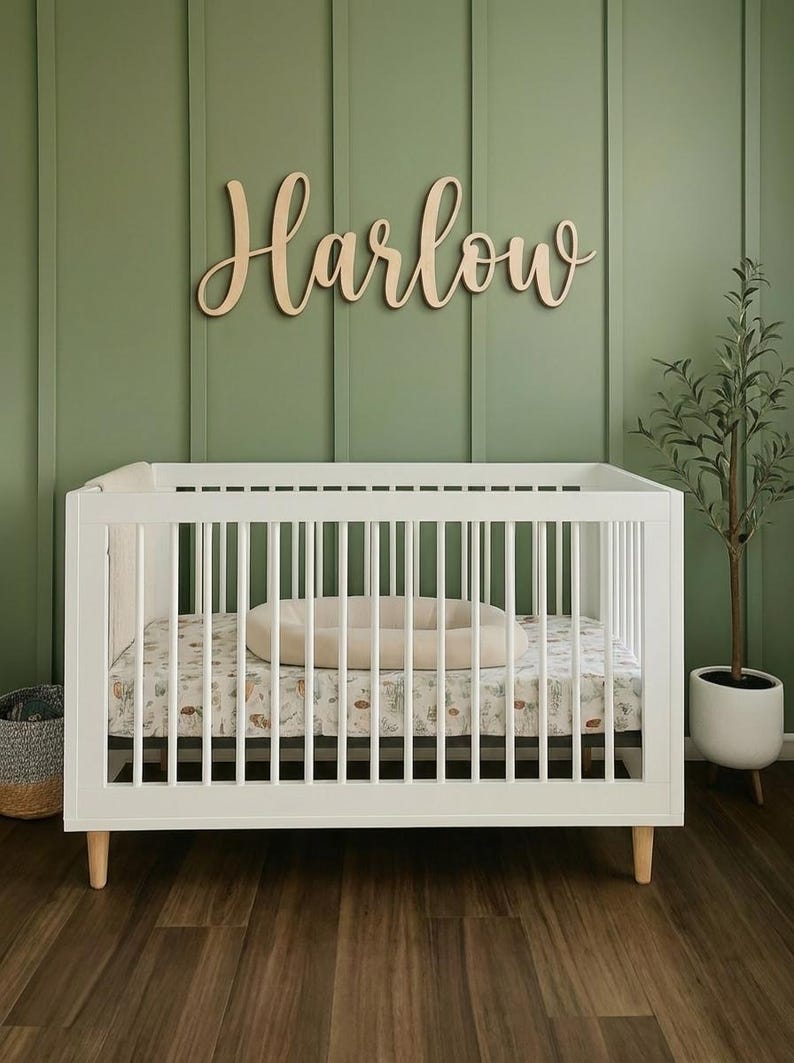 K&ouml;nnte beinhalten: Ein wei&szlig;es Babybett mit einer gemusterten Matratze und einem beigen Kissen. Das Wort "Harlow" ist in Holzschrift &uuml;ber dem Bett an einer gr&uuml;nen Wand angebracht. Rechts befindet sich eine kleine Topfpflanze.