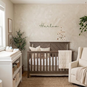 K&ouml;nnte beinhalten: Ein Kinderzimmer mit einem Holzbett, einer Wickelkommode und einem Schaukelstuhl. Das Bett ist aus braunem Holz und hat eine wei&szlig;e Matratze. An der Wand steht der Name "Harlow" in Schreibschrift. Ein Teppich in neutralen T&ouml;nen bedeckt den Boden.