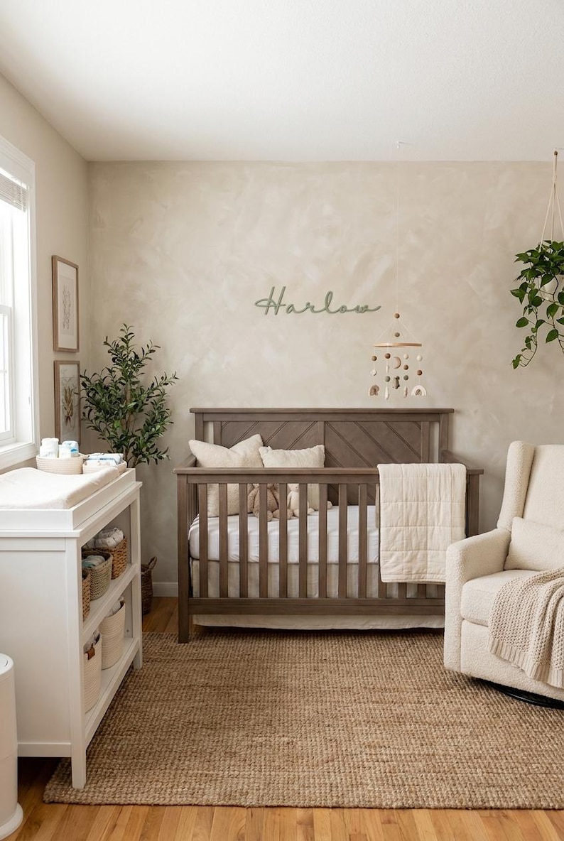 K&ouml;nnte beinhalten: Ein Kinderzimmer mit einem Holzbett, einer Wickelkommode und einem Schaukelstuhl. Das Bett ist aus braunem Holz und hat eine wei&szlig;e Matratze. An der Wand steht der Name "Harlow" in Schreibschrift. Ein Teppich in neutralen T&ouml;nen bedeckt den Boden.