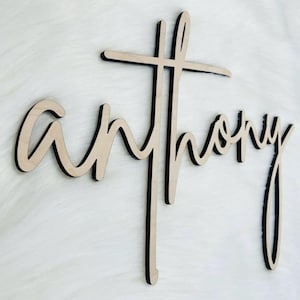 K&ouml;nnte beinhalten: Holzschild mit dem Namen "Anthony" in Schreibschrift, integriert in ein Kreuz. Das Schild ist aus hellem Holz mit schwarzen Kanten gefertigt, was einen geschichteten Effekt erzeugt. Es ist f&uuml;r die Pr&auml;sentation als Dekoration gedacht.