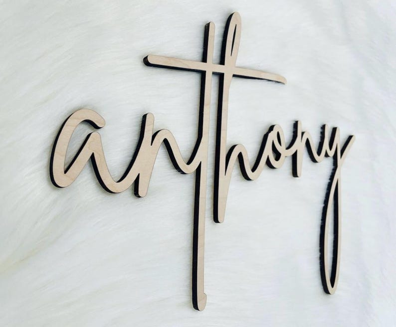 K&ouml;nnte beinhalten: Holzschild mit dem Namen "Anthony" in Schreibschrift, integriert in ein Kreuz. Das Schild ist aus hellem Holz mit schwarzen Kanten gefertigt, was einen geschichteten Effekt erzeugt. Es ist f&uuml;r die Pr&auml;sentation als Dekoration gedacht.