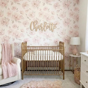 K&ouml;nnte beinhalten: Ein Kinderzimmer mit einem goldenen Kinderbett, einem wei&szlig;en Schaukelstuhl und einem rosa Teppich. Die Wand ist mit einer floralen Tapete mit rosa und wei&szlig;en Blumen bedeckt. Ein Holzschild mit der Aufschrift "Charlotte" ist an der Wand angebracht.