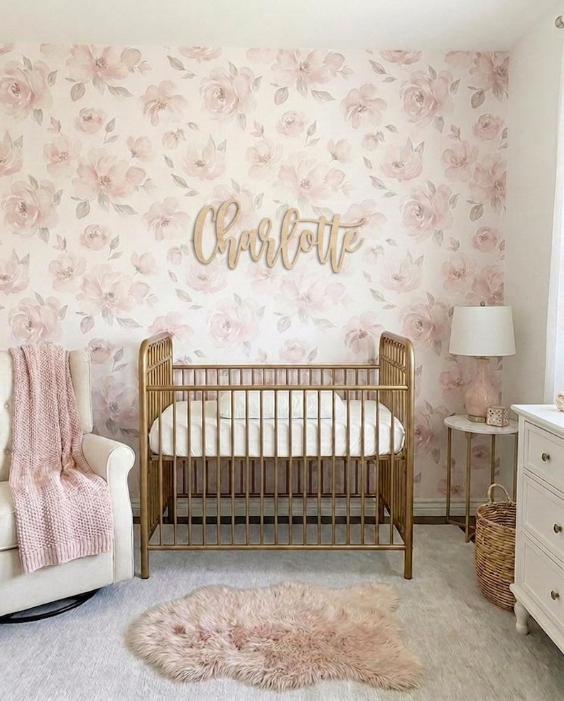 K&ouml;nnte beinhalten: Ein Kinderzimmer mit einem goldenen Kinderbett, einem wei&szlig;en Schaukelstuhl und einem rosa Teppich. Die Wand ist mit einer floralen Tapete mit rosa und wei&szlig;en Blumen bedeckt. Ein Holzschild mit der Aufschrift "Charlotte" ist an der Wand angebracht.