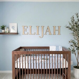 K&ouml;nnte beinhalten: Ein Holzbettchen mit Matratze und Bettw&auml;sche in einem Kinderzimmer. Der Name "ELIJAH" wird in Holzbuchstaben &uuml;ber dem Bettchen angezeigt. Ein kleines Regal mit B&uuml;chern und einem gerahmten Bild befindet sich an der Wand. Eine Topfpflanze befindet sich rechts.