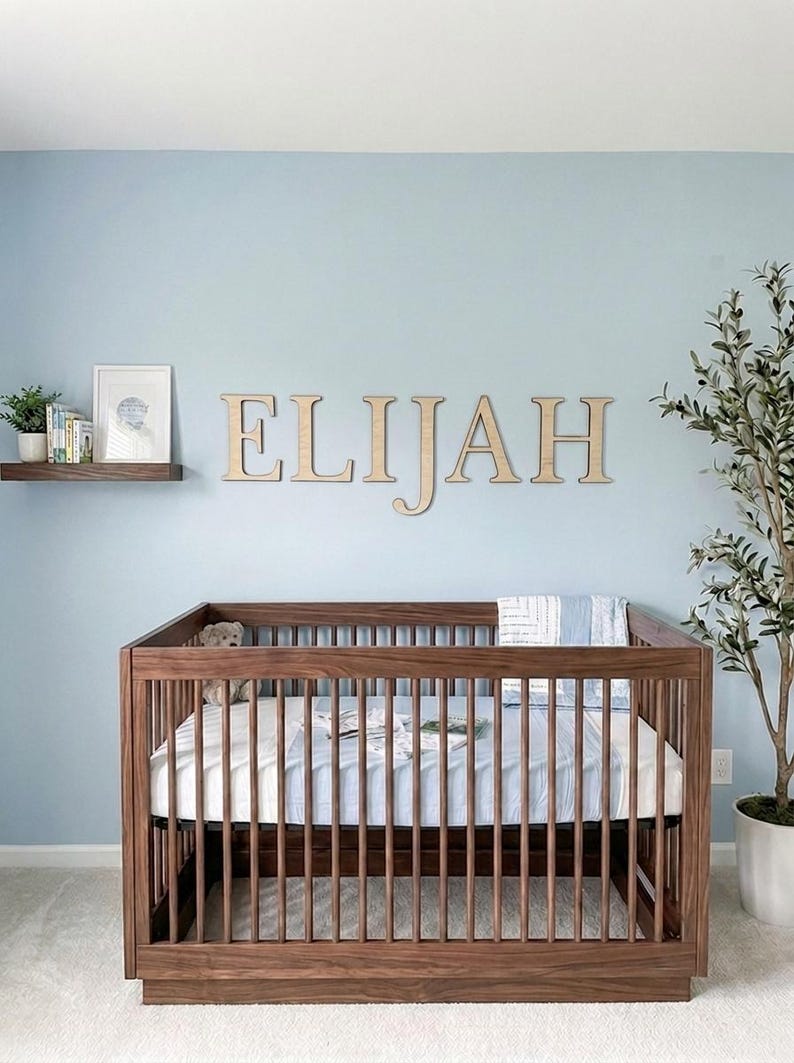 K&ouml;nnte beinhalten: Ein Holzbettchen mit Matratze und Bettw&auml;sche in einem Kinderzimmer. Der Name "ELIJAH" wird in Holzbuchstaben &uuml;ber dem Bettchen angezeigt. Ein kleines Regal mit B&uuml;chern und einem gerahmten Bild befindet sich an der Wand. Eine Topfpflanze befindet sich rechts.
