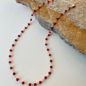 May include: A delicate gold chain necklace with small, round, red beads. The necklace is displayed on a rock.