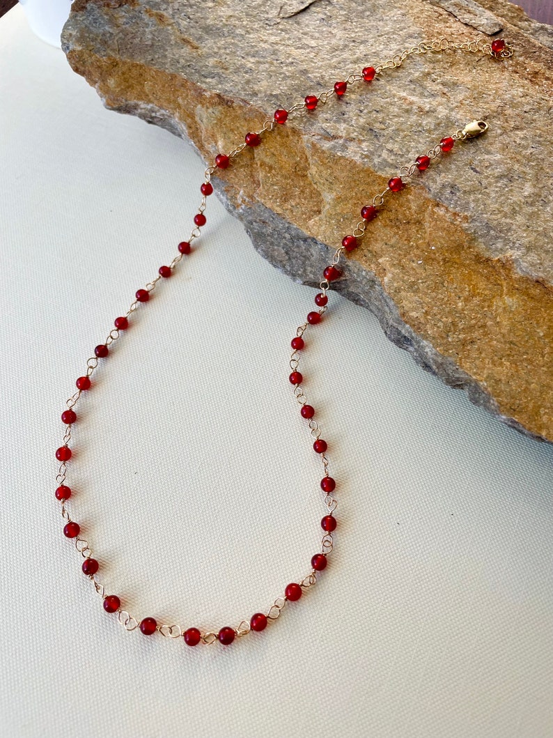 May include: A delicate gold chain necklace with small, round, red beads. The necklace is displayed on a rock.