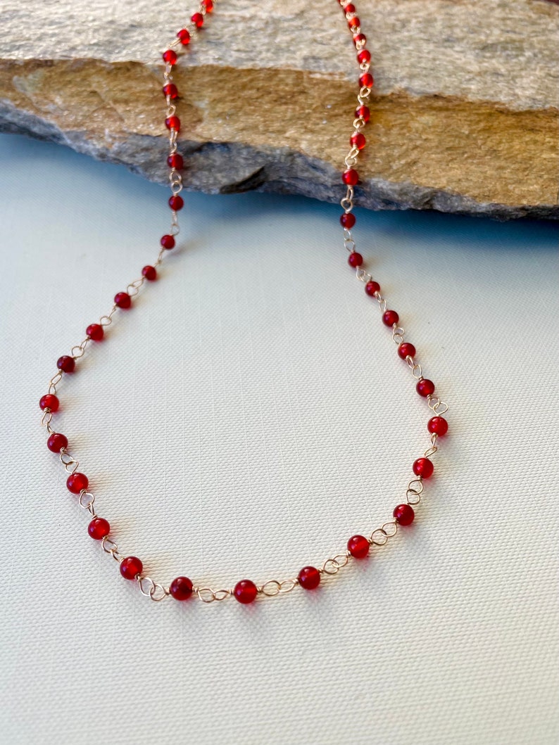 May include: A red carnelian gemstone necklace with a gold chain. The necklace is made of small, round beads and is approximately 18 inches long.