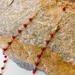 May include: A gold chain necklace with small red beads. The necklace is on a brown rock background.