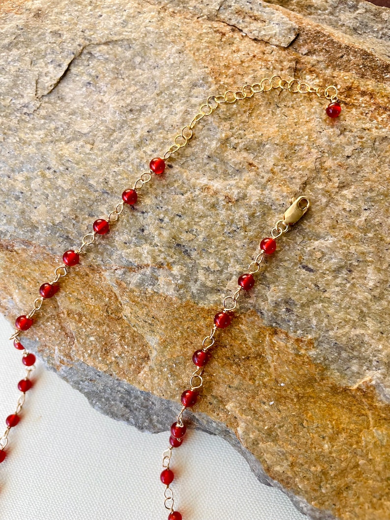 May include: A gold chain necklace with small red beads. The necklace is on a brown rock background.