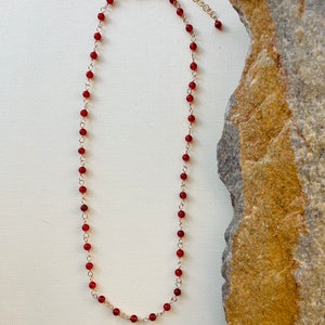 May include: A delicate gold chain necklace with small, round, red beads. The necklace is displayed on a white background with a rock to the right.