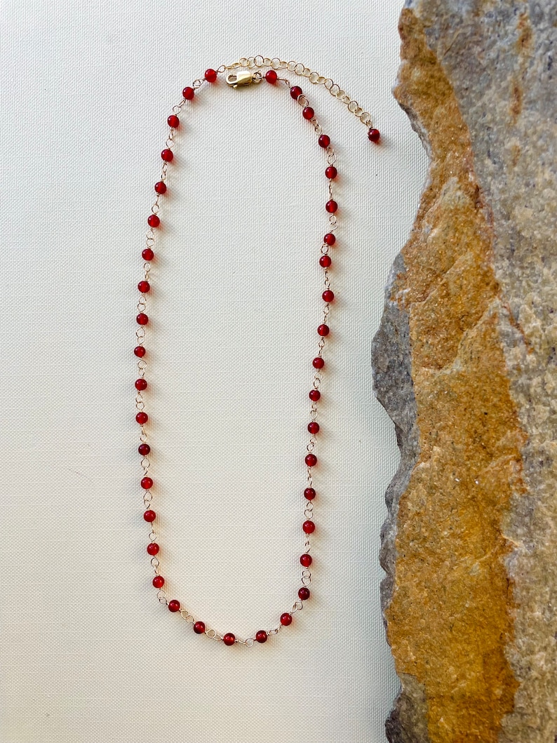 May include: A delicate gold chain necklace with small, round, red beads. The necklace is displayed on a white background with a rock to the right.