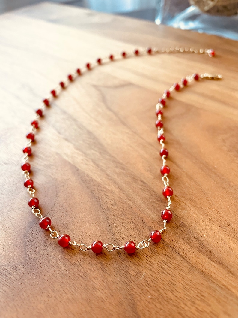 May include: A red carnelian gemstone necklace with gold chain links. The necklace is on a wooden surface.