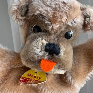 May include: A vintage plush dog puppet with brown and tan fur, black eyes, and an orange tongue. The puppet has a tag that reads "Mopsy" in yellow and red. The puppet's arms are outstretched.