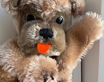 Puede incluir: Títere de perro de peluche marrón con ojos negros, una nariz bordada en negro y una lengua naranja. El pelaje del títere es suave y texturizado, con una expresión amigable y una pata levantada.