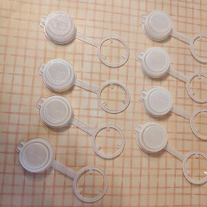 May include: Ten white plastic bottle stoppers with a ring handle. The stoppers are round and have a smooth surface.