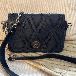 May include: Black leather crossbody bag with a diamond-quilted pattern. The bag features a gold chain strap and a gold logo emblem on the front. The adjustable strap is black leather. The bag is sitting on a beige surface.