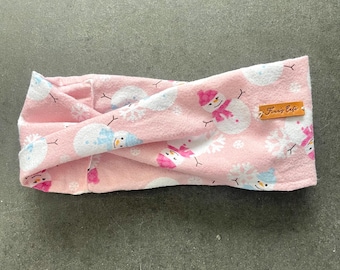 May include: A pink fleece headband with a repeating pattern of snowmen, snowflakes, and clouds. The snowmen wear pink hats and scarves. A small brown tag with the text "Farris Cafe" is sewn on the side. This winter accessory is ideal for keeping warm.