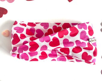 May include: A white headband with a pink and red heart pattern. The headband is made of fabric and has a small tag that says "Fawn & Co."