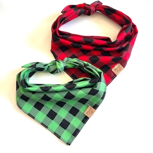 May include: Two dog bandanas in a buffalo plaid pattern. One is red and black, the other is green and black. Each bandana has a small brown tag that reads "Tiny Cafe". The bandanas are knotted at the top.