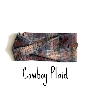 May include: A twisted headband featuring a multi-coloured plaid pattern in shades of brown, blue, and tan. A small brown leather tag with the text Kari's Cafe is attached to the side. The text Cowboy Plaid is printed below the headband.