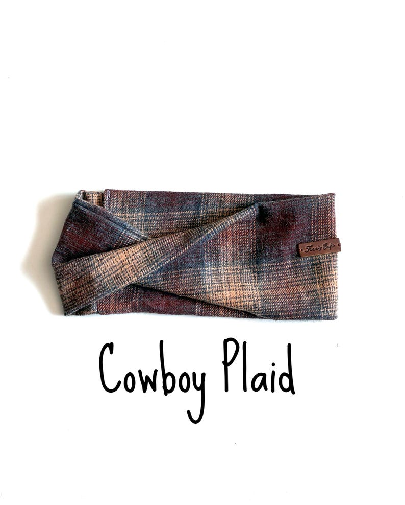 May include: A twisted headband featuring a multi-coloured plaid pattern in shades of brown, blue, and tan. A small brown leather tag with the text Kari's Cafe is attached to the side. The text Cowboy Plaid is printed below the headband.