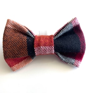 May include: A bow tie hair accessory featuring a multi-coloured plaid pattern in shades of orange, red, black, and white. The fabric has a visible woven texture, and a small clear plastic hair comb is attached to the centre back of the bow.