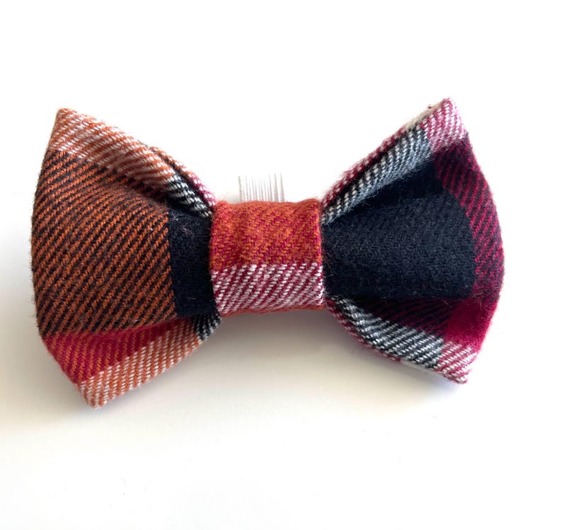May include: A bow tie hair accessory featuring a multi-coloured plaid pattern in shades of orange, red, black, and white. The fabric has a visible woven texture, and a small clear plastic hair comb is attached to the centre back of the bow.