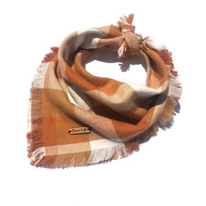 May include: A triangular pet bandana featuring a burnt orange and cream plaid pattern with frayed edges. The fabric is tied in a knot at the top and includes a small rectangular wooden tag attached to the front with the text Finn's Cafe engraved.
