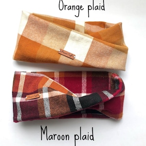 May include: Two fabric headbands with a twisted design. The top headband features an orange and cream plaid pattern, while the bottom headband displays a maroon, orange, white, and black plaid pattern. Each headband has a small brown leather tag labelled Finn's Cafe.