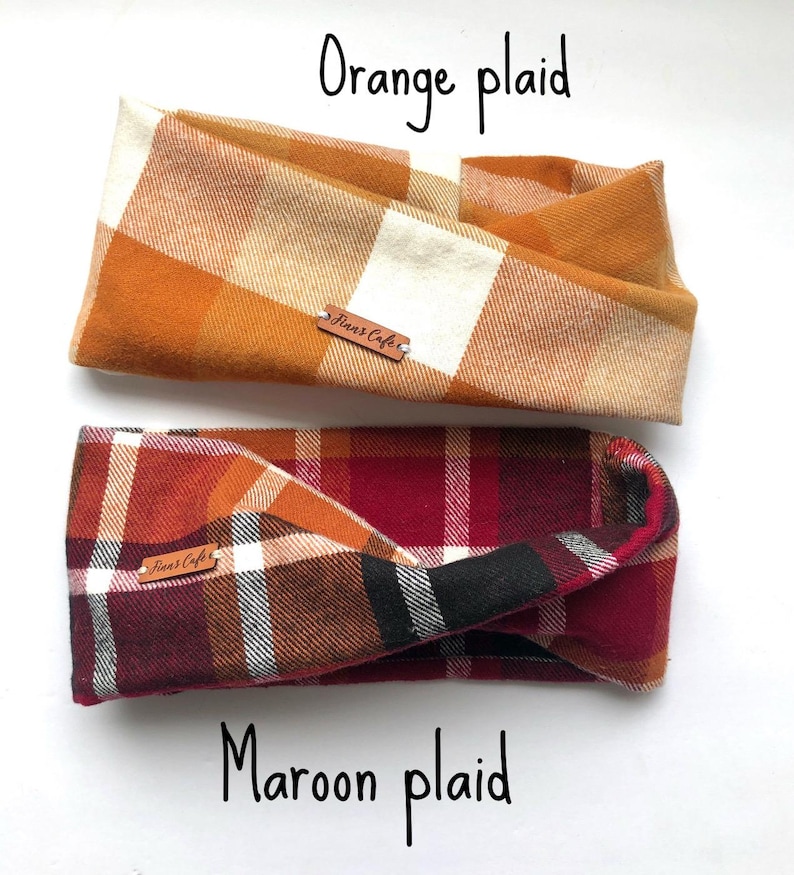 May include: Two fabric headbands with a twisted design. The top headband features an orange and cream plaid pattern, while the bottom headband displays a maroon, orange, white, and black plaid pattern. Each headband has a small brown leather tag labelled Finn's Cafe.